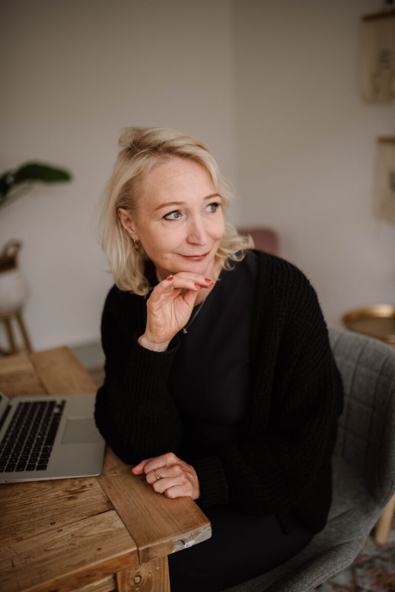 Relatiecoach Anne de Jong zit aan tafel met laptop en kijkt nadenkend opzij, symbool voor reflectie en inzicht in relaties